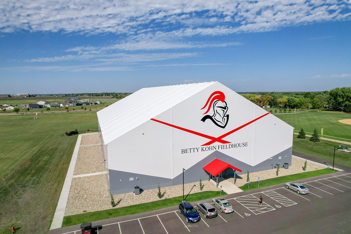 Exterior view of the Betty Kohn Fieldhouse, a bright white hybrid fabric structure with bold red graphics and a knight logo, surrounded by green fields and parking.