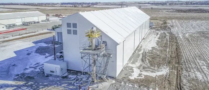 A steel I‑beam rigid frame under construction with corrugated concrete lower walls and a clear tension fabric roof being placed above—a durable industrial structure set on gravel under an overcast sky.